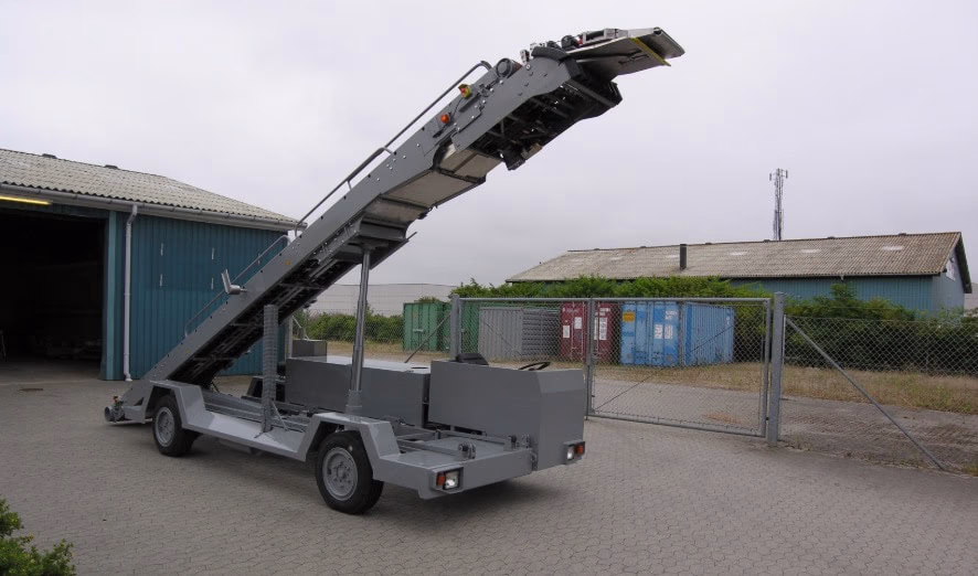 A belt loader is parked on a paved surface near a chain-link fence, with industrial buildings and shipping containers in the background.